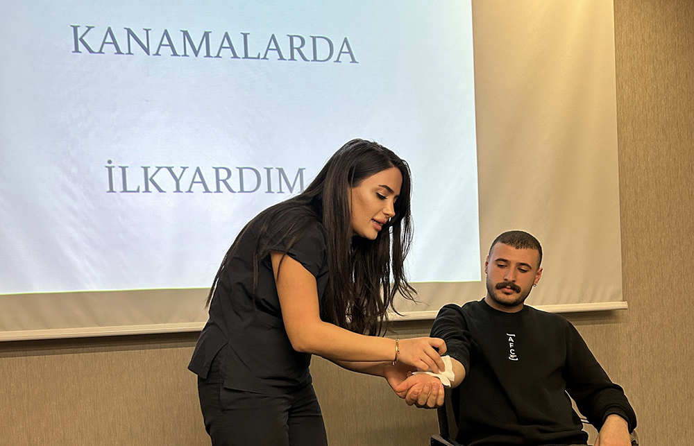 Kanama Durumlarında İlkyardım konulu bir eğitimde, bir ilk yardımcı kadın eğitmen erkek bir katılımcının koluna bandaj uygulayarak pratik gösterim yapıyor.