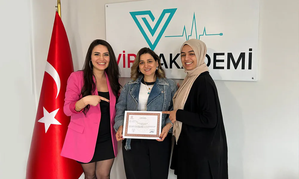 Vira Akademi logolu tabela önünde, bir Türk bayrağı yanında duran üç kadın. Ortadaki kadın, gülümseyerek bir sertifika tutarken, sağdaki ve soldaki kadınlar parmaklarıyla sertifikayı işaret ederek başarıyı kutluyorlar.